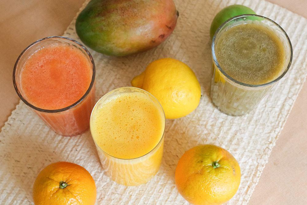 Drei verschiedene gesunde Smoothies in Gläsern neben Mango und Orangen