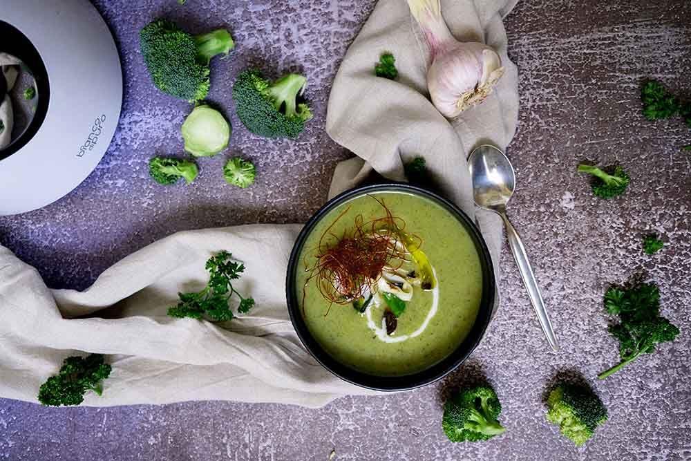 Dunkle Schale mit grüner Brokkoli-Cremesuppe, garniert mit Chili-Fäden und Rahm, umgeben von frischem Brokkoli und Lauch.
