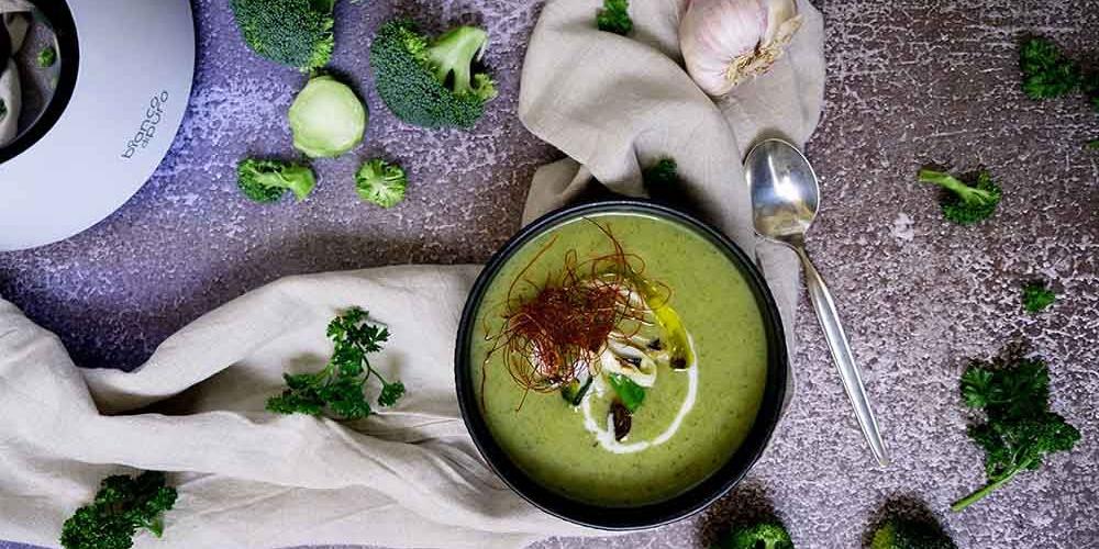 Dunkle Schale mit grüner Brokkoli-Cremesuppe, garniert mit Chili-Fäden und Rahm, umgeben von frischem Brokkoli und Lauch.