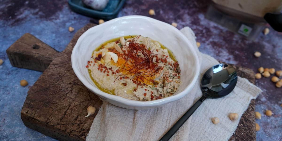 Nahaufnahme von hausgemachtem Hummus in einer weissen Schale mit Gewürzen und Olivenöl auf einem Holzbrett.