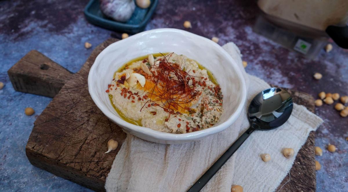 Nahaufnahme von hausgemachtem Hummus in einer weissen Schale mit Gewürzen und Olivenöl auf einem Holzbrett.