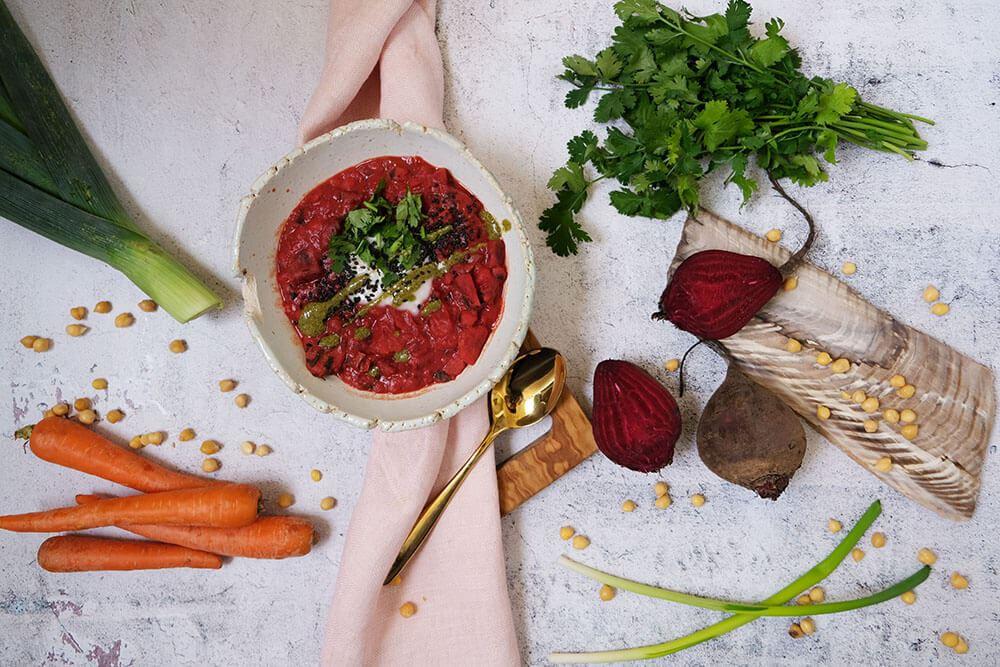 Eine Schale mit leuchtend roter Kichererbsen-Suppe, garniert mit frischem Koriander, Karotten und Lauch im Hintergrund.